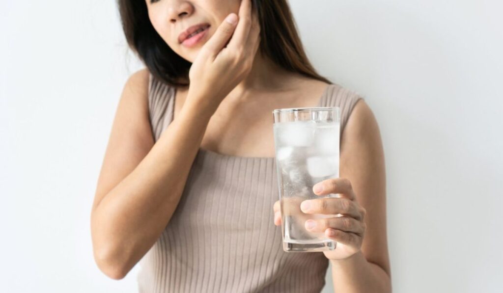 Young Asian woman with sensitive teeth and hand holding glass of cold water with ice