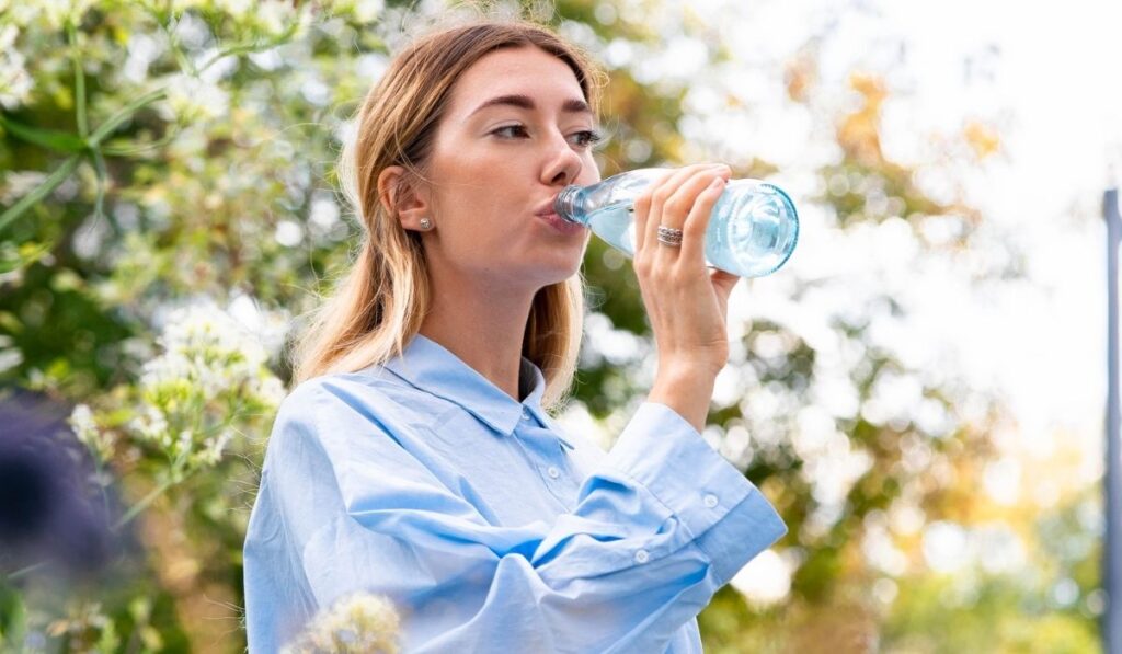 Young and attractive woman drinking sparkling water from glass eco biodegradable bottle at modern park