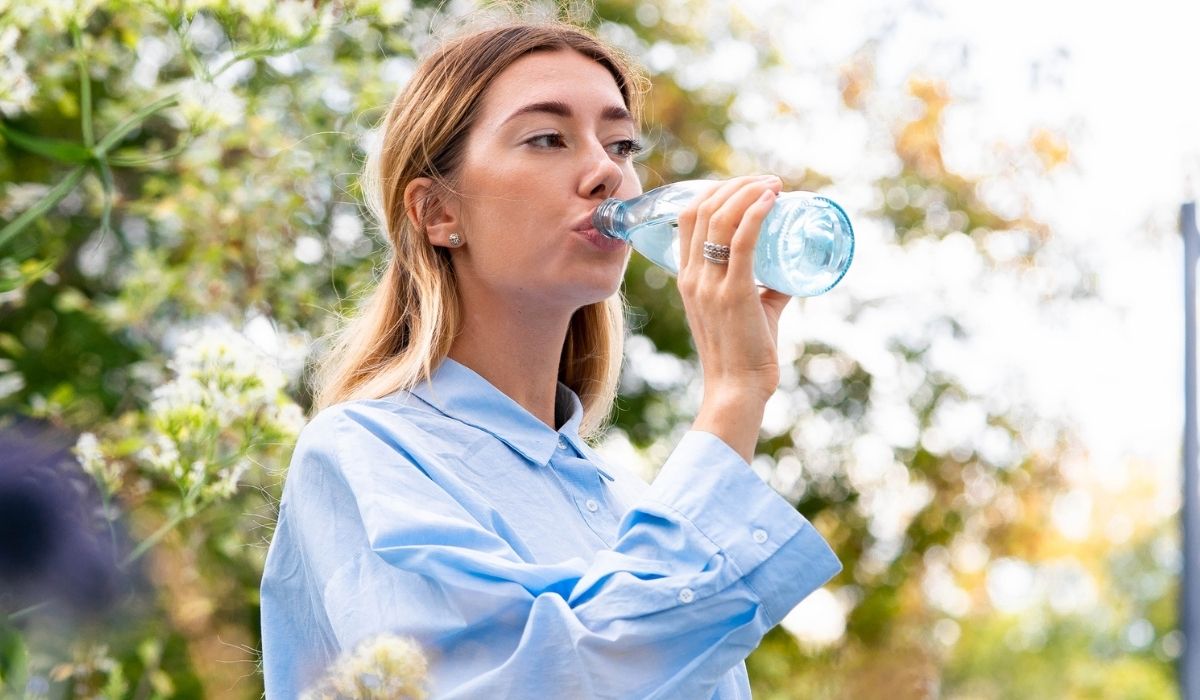 Young and attractive woman drinking sparkling water from glass eco biodegradable bottle at modern park