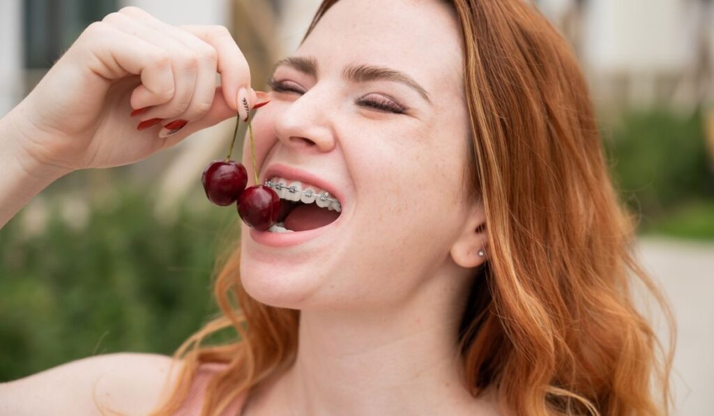 Beautiful young red-haired woman with braces on her teeth eats sweet cherries in the summer outdoors 