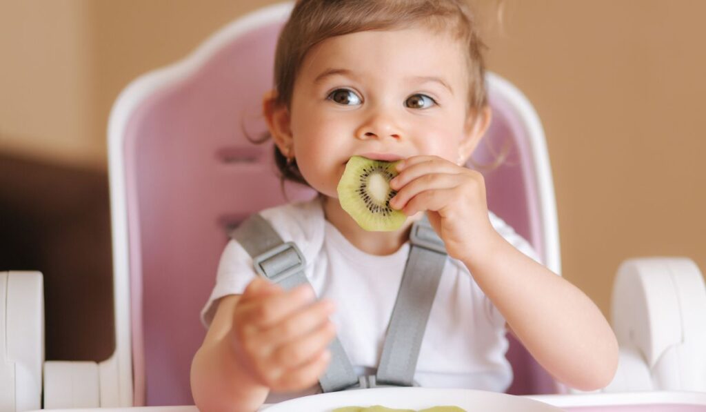 Portrait of happy little kid eating kiwi in high chair