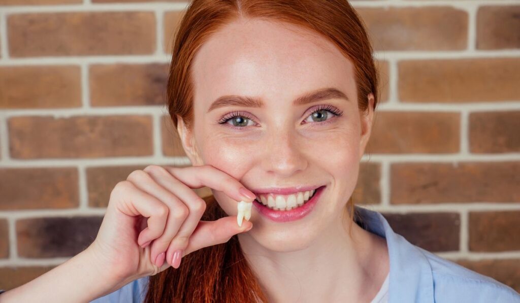 Redhaired ginger female with snow-white smile holding white wisdom tooth after surgery removal of a tooth
