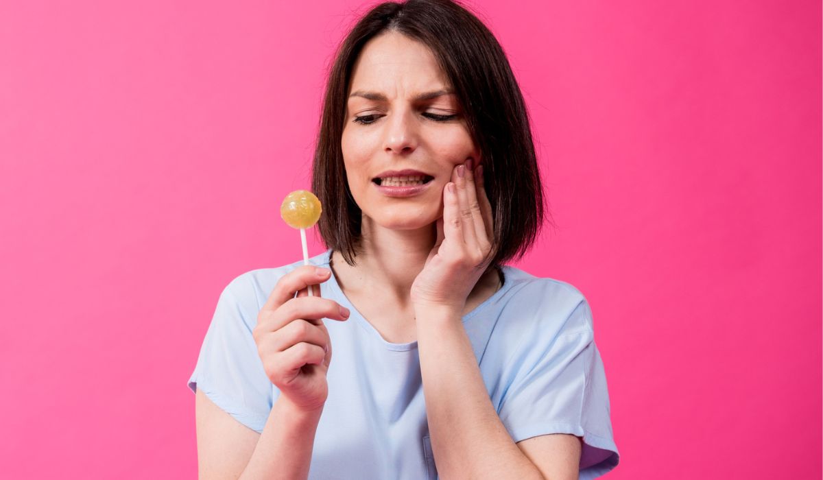 Young woman with sensitive teeth eating sweet lollipop on color background